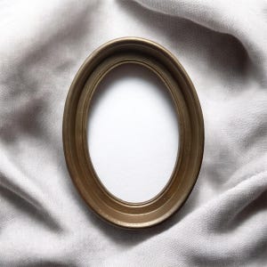 May include: A gold-toned, oval picture frame with a minimalist design. The frame is empty, with a white interior, and rests on a textured, light grey fabric. The frame is a decorative item.