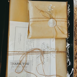 May include: A gift box with brown paper wrapping, tied with twine, and adorned with sprigs of white flowers. The box contains a certificate of authenticity and a thank you card. The text "Thank You for your order" is visible.
