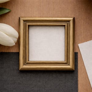 May include: A square, gold-colored picture frame with a white interior. The frame is set on a wooden surface, with a white tulip and dark gray fabric in the background. The frame is empty, ready for a photo or artwork.