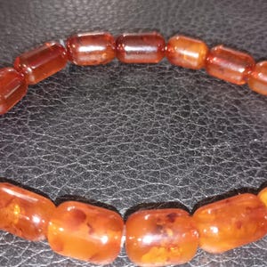 May include: A bracelet made of amber-coloured, barrel-shaped beads. The beads are strung together, forming a circular shape. The amber beads have a warm, reddish-brown hue and a polished appearance. The bracelet is displayed on a dark, textured surface.