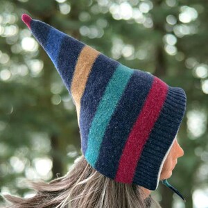 Peut inclure: Un bonnet en tricot rayé avec un long sommet pointu. Le chapeau présente des bandes bleu marine, beige, turquoise et rouge. Le bout du chapeau est une petite pointe rose. Le chapeau est porté par une personne aux longs cheveux châtain clair.