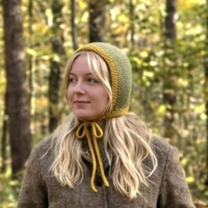 Op de afbeelding: Een handgemaakte gehaakte muts in olijfgroen met een mosterdgele rand en strikjes. De muts wordt gedragen door een persoon met blond haar, en de achtergrond is een wazig zicht op een bos met herfstbladeren.