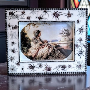 May include: A rectangular picture frame with a white background, decorated with black spiders and webs. The frame has a black beaded border. The framed image depicts a woman and child in a landscape setting.