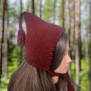 Puede incluir: Un gorro de punto burdeos con una larga corona puntiaguda y un borlón. El gorro tiene un borde de ganchillo y una larga corbata que se puede atar debajo de la barbilla.