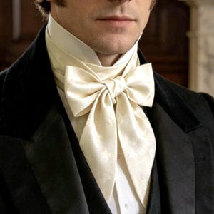 May include: A cream-colored silk cravat featuring a subtle star pattern, tied in a formal bow at the neck. The cravat is worn over a crisp white collared shirt and underneath a black velvet jacket with a high collar and dark lapels.