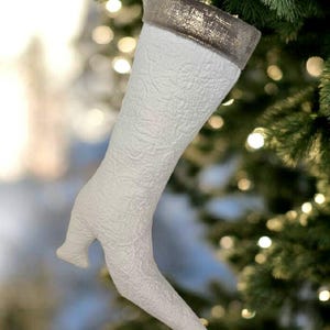Op de afbeelding: Een witte kerstsok in de vorm van een high-heeled laars, hangend aan een kerstboom. De sok heeft een textuurpatroon en een metallic gouden manchet. De hak is een kleine, gebogen vorm.