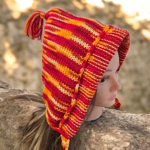 Puede incluir: Un gorro tejido a mano con rayas rojas, naranjas y amarillas. El gorro tiene una parte superior puntiaguda, orejeras y una borla. El gorro se muestra en una cabeza de maniquí. Un accesorio de invierno.