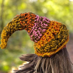 May include: A handmade crochet hat in a mix of colours including yellow, orange, pink, and green. The hat has a curved, pointed shape, and is made with a thick yarn. The hat is shown on a person's head, with long brown hair visible.