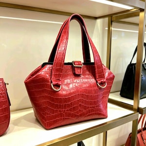 May include: A red handbag with a textured crocodile-embossed pattern, featuring two top handles, a central metal buckle closure, and gold-toned hardware rings at the base of the handles. The bag has a structured, wide-bottomed silhouette.