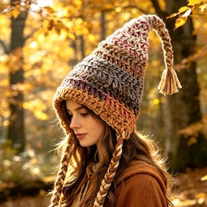 May include: A chunky knit beanie with a tall, pointed crown that curves downwards at the tip, finished with a braided tassel. The hat features horizontal stripes in variegated shades of brown, cream, and rust, with two long braided side ties.