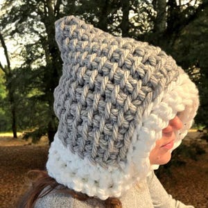 Puede incluir: Un gorro de ganchillo hecho a mano en gris y blanco. El gorro presenta una parte superior puntiaguda y un borde blanco ancho. La sección gris tiene una apariencia texturizada y tejida. El gorro está diseñado para ser cálido y cómodo.