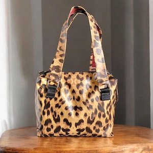 May include: A small leopard print tote bag with black straps and buckles. The bag has a red lining.