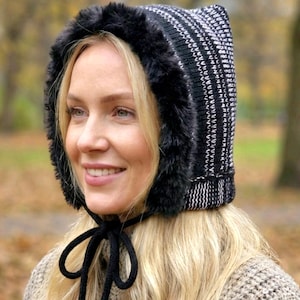 Puede incluir: Un gorro de invierno de punto blanco y negro con ribete de piel sintética. El gorro tiene un patrón de rayas y un cierre de lazo negro. Este gorro está diseñado para mantener la cabeza y las orejas calientes en climas fríos.