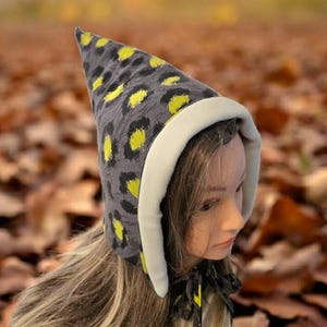 Puede incluir: Un gorro de duende con estampado de leopardo gris y amarillo con ribete y lazos blancos. El gorro está hecho de tela suave y tiene una parte superior puntiaguda. El fondo está borroso con hojas de otoño.