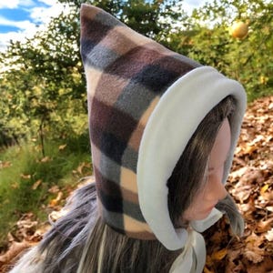 Puede incluir: Una capucha de forro polar a cuadros en tonos marrones, grises y crema, con una parte superior puntiaguda y un ribete de forro polar blanco alrededor de la cara. Una cinta blanca está atada al cuello. La capucha está diseñada para ser usada.