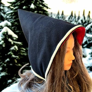 Könnte beinhalten: Ein schwarzer Kapuzenmantel mit spitzer Spitze, rot gefüttert. Die Kapuze ist mit einer weißen und goldenen Borte verziert. Der Hintergrund zeigt eine verschneite Waldszene.