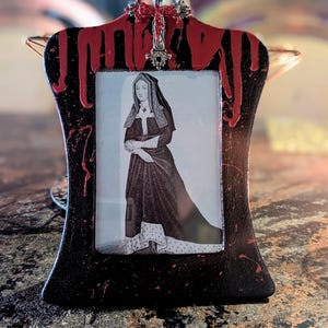 May include: A black picture frame with red paint dripping down the sides. The frame has a silver cross at the top and a black and white image of a woman in a long dress inside.