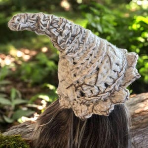 Può includere: Un cappello da gnomo all'uncinetto fatto a mano in crema e marrone. Il cappello presenta una parte superiore a punta e un bordo smerlato. Il filato ha un aspetto strutturato, con punti visibili e un mix di fili marroni chiari e scuri. Il cappello è progettato per essere indossato.