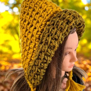 Puede incluir: Un gorro de ganchillo hecho a mano en tonos dorados y verde oliva. El gorro presenta un patr&oacute;n de puntadas texturizadas y una capucha puntiaguda. Una borla cuelga de un lado. El gorro se exhibe en una cabeza de maniqu&iacute;.