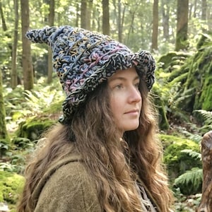 Puede incluir: Un sombrero de bruja de ganchillo multicolor hecho a mano con una corona puntiaguda y un ala ancha. El sombrero presenta una mezcla de hilo negro, azul, amarillo y rosa. El sombrero se usa al aire libre en un entorno forestal.