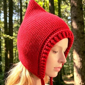 May include: A bright red knitted bonnet with a pointed top and a textured ribbed border around the face opening. The headwear features two long, matching red knitted ties hanging down from the sides, crafted with a consistent vertical stitch pattern.