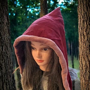 Puede incluir: Una capa con capucha burdeos con una punta, ribeteada con una tela rosa-marrón más clara. La capucha se muestra en una cabeza de maniquí con cabello largo y oscuro, contra un fondo borroso de bosque.
