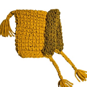 Puede incluir: Un bolso de ganchillo hecho a mano en amarillo mostaza y verde oliva. El bolso tiene forma rectangular con una solapa y dos largas borlas. La puntada de ganchillo crea una superficie texturizada. Las borlas son de color amarillo mostaza a juego.