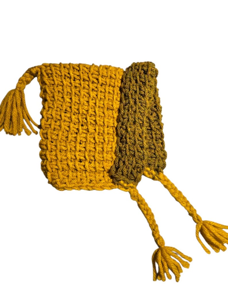 Puede incluir: Un bolso de ganchillo hecho a mano en amarillo mostaza y verde oliva. El bolso tiene forma rectangular con una solapa y dos largas borlas. La puntada de ganchillo crea una superficie texturizada. Las borlas son de color amarillo mostaza a juego.