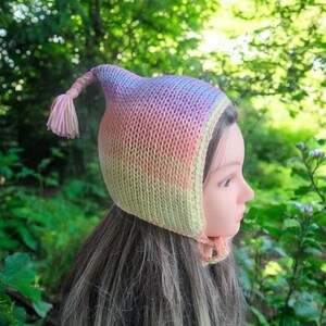 May include: A rainbow ombre knitted baby bonnet with a long tassel. The bonnet is made of soft yarn and has a pointed top.