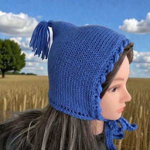 Op de afbeelding: Een blauwe gebreide muts met een kwast en striksluitingen. De muts heeft een geschulpte rand en is gemaakt van zacht, getextureerd garen. De muts wordt getoond op een mannequinhoofd.