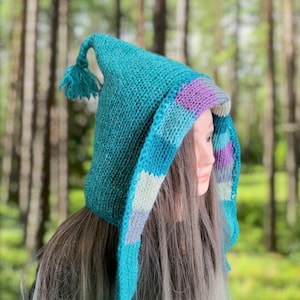 Può includere: Un cappello di lana color turchese con una corona a punta e una lunga sciarpa fluida. Il cappello presenta un motivo a righe colorate sul lato, con sfumature di viola, blu e bianco.