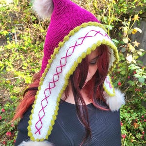 Puede incluir: Un gorro de punto rosa con ribete blanco y verde. El gorro tiene un pompón en la parte superior y un ribete de piel blanca alrededor del borde.