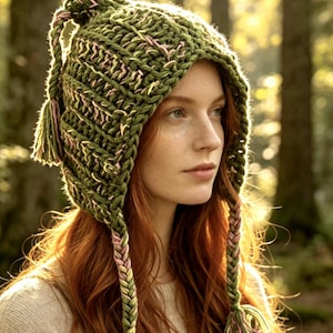 May include: A chunky knit olive green hat featuring a pointed top with a tassel, two long braided side ties ending in tassels, and subtle interwoven strands of light pink and yellow yarn throughout the thick, textured crochet construction.