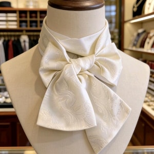 May include: Ivory ascot tie with a paisley pattern, displayed on a mannequin. The tie is knotted in a bow shape, showcasing the intricate design. The background suggests a retail setting, with shelves and clothing visible.