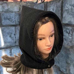 Puede incluir: Una capa con capucha negra hecha de una tela oscura y suave. La capucha es puntiaguda y tiene un cuello redondo que se abrocha con un botón. La capa se muestra en una cabeza de maniquí con cabello castaño largo.