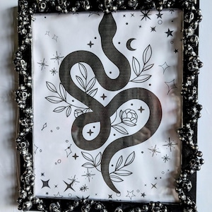 Puede incluir: Impresión artística enmarcada en negro con una ilustración de serpiente negra, elementos florales y celestiales. El marco negro está adornado con calaveras plateadas. La obra de arte está sobre un fondo blanco.