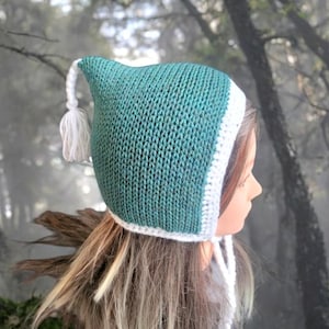Puede incluir: Un gorro de punto de color turquesa con ribete de crochet blanco y un borla blanca. El gorro tiene una corona puntiaguda y una larga corbata fluida.