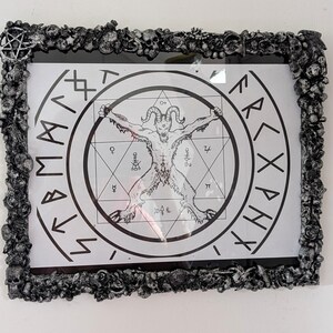 May include: A rectangular wall art piece featuring a black and white illustration of a horned figure inside a circular frame with runic symbols and geometric shapes. The frame is textured, dark grey, and decorated with small skulls and organic forms.