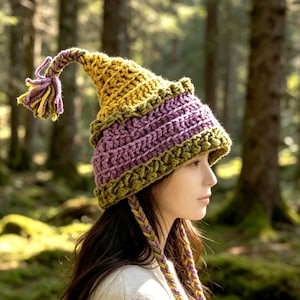 May include: A crocheted hat featuring a pointed top with a tassel, constructed from alternating bands of mustard yellow and purple yarn. The hat includes a textured olive green border at the base and two long braided ties hanging from the sides.