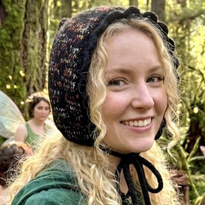 May include: A black bonnet with a multi-coloured speckled pattern and a scalloped edge. The bonnet is tied with black cords. The person in the image is wearing a green velvet top. The background is a forest.