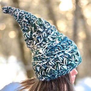 Puede incluir: Un gorro puntiagudo hecho a mano en tonos turquesa, blanco y azul marino. El gorro está tejido a ganchillo con un punto texturizado, con una punta larga y curvada. El gorro está diseñado para ser usado.