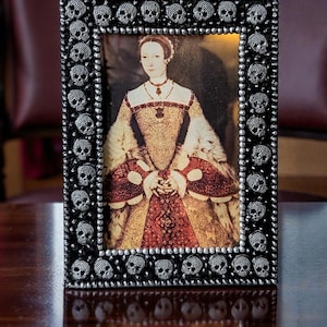 Könnte beinhalten: Ein rechteckiger, schwarzer Bilderrahmen, verziert mit kleinen silbernen Totenköpfen. Er enthält ein Porträt einer Frau in einem historischen Kleid. Die Frau trägt ein goldenes und rotes Kleid mit weißem Pelzkragen.