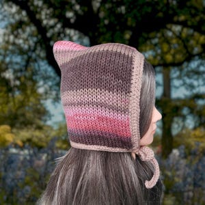 May include: A knitted hooded hat with a striped pattern in shades of pink, brown, and beige. The hat has a long tie that can be tied under the chin.