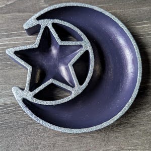Op de afbeelding: Een donkerpaarse sieradenschaal in de vorm van een halve maan met een ster in het midden. De randen van de schaal en de ster zijn omrand met zilveren glitter. De schaal staat op een houten oppervlak.