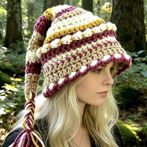 May include: A long, pointed crochet hat featuring horizontal stripes in maroon, cream, and mustard yellow. The hat has a textured, chunky knit pattern with a scalloped edge and a long, braided tail ending in loose yarn tassels of the same colours.