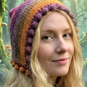 Puede incluir: Gorro tejido a mano con colores vibrantes: morado, naranja y marrón. Presenta un borde con pompón y una parte superior puntiaguda. La persona que lo lleva tiene el pelo rubio, sobre un fondo de bosque borroso.