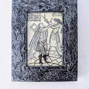 May include: A framed print in black and white showing a barber trimming a man's beard. The dark grey frame has a floral relief design. The print features two figures in historical attire, one holding scissors.