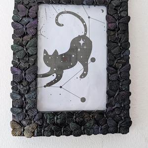 May include: Black rectangular picture frame decorated with butterflies. The frame holds a black and white illustration of a cat with stars and constellations. The butterflies on the frame are in shades of purple, green, and black.
