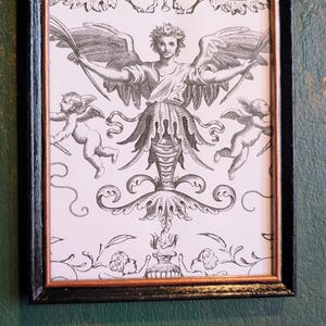 May include: Black framed print featuring a detailed black and white illustration of an angel with outstretched wings, cherubs, and ornate floral elements. A small cherub sculpture adorns the top of the frame. The artwork is displayed against a green wall.
