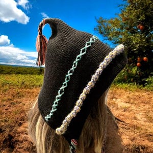 May include: A black knit hat with a pointed top and a decorative tassel. The hat features a light blue and white stitched design along the side and a colorful trim. The background shows a blue sky and a field.